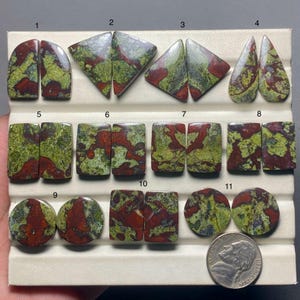 Könnte beinhalten: Eine Sammlung von elf Paaren polierter Edelsteine in verschiedenen Formen, darunter Tropfen und Rechtecke. Die Steine weisen ein gesprenkeltes Muster aus roten, grünen und braunen Farbtönen auf. Eine Münze dient als Größenvergleich.