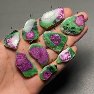 May include: Nine rough ruby in zoisite stones are displayed. Each stone has a striking contrast of deep magenta ruby inclusions within a green zoisite matrix. The stones' sizes and shapes vary, highlighting natural textures and patterns.