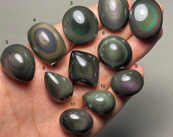 May include: A collection of eleven polished obsidian gemstones in various shapes, including oval, teardrop, and rounded forms. The stones display iridescent bands of color, with hues of green, purple, and gold, against a dark, reflective surface.