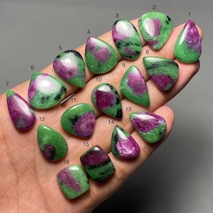 May include: Sixteen ruby zoisite gemstones in diverse shapes, such as teardrop and oval. The stones exhibit a mix of vibrant green and deep magenta hues, with natural patterns. The gemstones are presented against a neutral backdrop. Measurements are in centimeters.