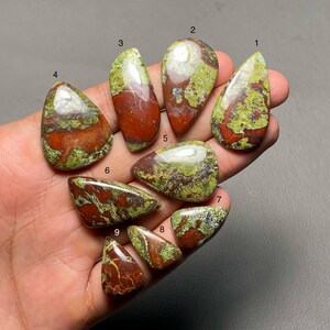May include: Nine polished gemstones of varying shapes and sizes, held in a hand. The stones display a mix of red, green, and brown hues. Each stone has a unique pattern, ideal for jewellery making or collecting.