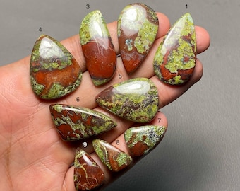 May include: A collection of nine polished gemstones in various shapes, held in a hand. The stones feature a mix of red, green, and brown colors. Each stone has a unique pattern and shape, suitable for jewelry or collecting.