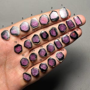 Könnte beinhalten: Eine Sammlung von 27 rohen Turmalin-Edelsteinen, jede mit einer einzigartigen Form und Farbe. Die Steine reichen von Rosa bis zu tiefem Magenta, mit schwarzen Rändern. Die Edelsteine sind von 1 bis 27 nummeriert.