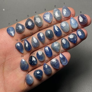 Puede incluir: Una colección de veintinueve piedras preciosas de zafiro azul en varias formas, incluyendo ovaladas, de pera y de lágrima. Cada piedra está facetada, mostrando una gama de tonos azules. Las piedras preciosas están numeradas del 1 al 29.