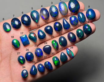 May include: A collection of 34 black opal gemstones, each with unique patterns and colors. The opals vary in shape, including oval, teardrop, and rectangular cuts. The gemstones display flashes of green, blue, and other colors.