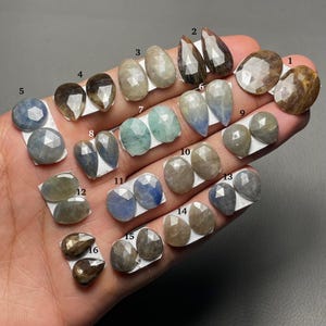 May include: Sixteen faceted gemstones in diverse shapes and colours, including oval, teardrop, and round cuts. The gemstones vary in colour from blue and green to brown and grey. Each stone is displayed on a white backing.