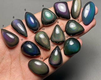 May include: A collection of fourteen polished rainbow obsidian gemstones in various shapes, including teardrop, oval, and rounded forms. The stones display iridescent colors of blue, green, purple, and gold, with a dark, glossy base. The gemstones are held in a hand.