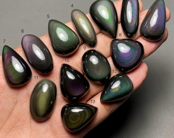 May include: A collection of thirteen polished obsidian gemstones in various shapes, held in a hand. The stones display iridescent colors including green, purple, and gold, with a glossy, reflective surface. Each stone is unique in its pattern and color distribution.