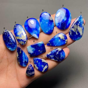 May include: A collection of twelve polished lapis lazuli gemstones in various shapes, including oval, teardrop, and rectangular. The stones display a deep blue color with streaks and flecks of gold and white, showcasing natural patterns.