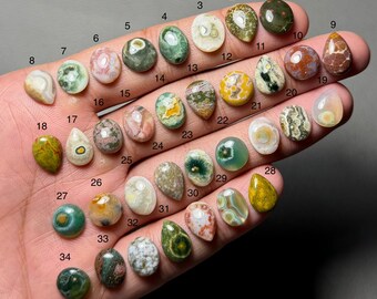 May include: A selection of 34 polished gemstones in diverse shapes and colours, including green, brown, yellow, and white. Each stone is numbered and displayed on a hand, showcasing the varied patterns and textures of the natural stones.