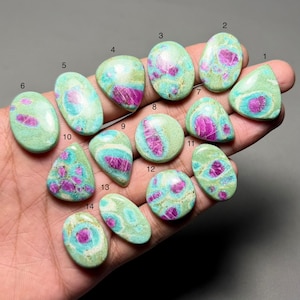 May include: A collection of fourteen polished ruby zoisite gemstones. Each stone features a unique pattern of vibrant pink and green hues. The gemstones vary in shape, including oval, teardrop, and round, showcasing natural variations in color and texture.