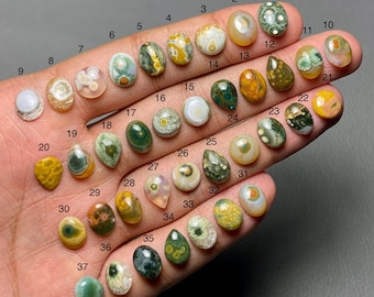 May include: A selection of 37 polished gemstones in diverse shapes and colours, including green, yellow, brown, and white. The gemstones are numbered and displayed on a hand, highlighting their unique patterns and textures.