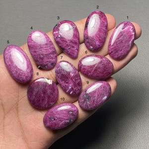 以下が含まれることがあります： さまざまな形をした、楕円形や涙滴型を含む、10個の研磨されたルビーカボションのコレクション。宝石は、微妙な内部パターンと暗いインクルージョンを持つ、深いピンクがかった赤色を示しています。カボションは手の上に表示されています。