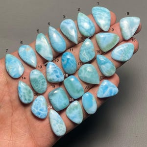 May include: A collection of twenty-four polished larimar gemstones in various shapes and sizes, ranging from teardrop to oval. The stones display a range of blue and white hues, with some exhibiting a marbled appearance. The gemstones are held in a hand.