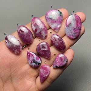 May include: Ten polished ruby cabochons in diverse shapes, including oval, teardrop, and pear. The gemstones exhibit deep pink and white hues, with some showing black inclusions. The stones are displayed on a neutral background.