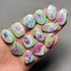 Puede incluir: Una colección de trece piedras preciosas pulidas, cada una con una forma única. Las piedras presentan una matriz verde claro con inclusiones rosas y magenta vibrantes. Las piedras preciosas se sostienen en una mano, mostrando su variedad.