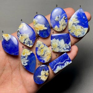May include: A collection of ten polished gemstones in various shapes, including teardrop, oval, and rectangular. The stones feature a deep blue base color with swirling patterns of creamy yellow and white, creating a unique, natural appearance.
