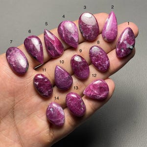 Könnte beinhalten: Eine Sammlung von vierzehn polierten Rubin-Edelsteinen in verschiedenen Formen, darunter oval, tropfenförmig und spitz. Die Edelsteine zeigen einen tiefen, satten rosaroten Farbton, einige mit weißen Einschlüssen. Die Steine werden auf einer Hand präsentiert.