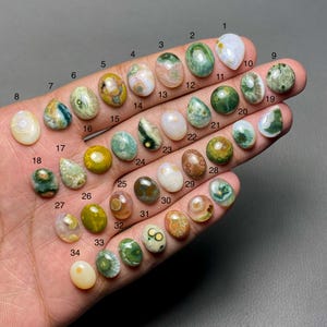May include: A selection of thirty-four polished gemstones in diverse shapes and colours, including green, brown, white, and yellow. Each stone is numbered and displayed on a hand, showcasing unique patterns and textures. The gemstones vary in size and shape.
