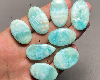 May include: A collection of eight polished gemstones in various shapes, held in a hand. The stones display a range of blue and white hues, with unique patterns and textures. The gemstones are oval, teardrop, and rounded shapes.