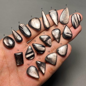 Puede incluir: Una colección de dieciocho piedras preciosas pulidas de varias formas, incluyendo ovaladas, en forma de lágrima y rectangulares. Las piedras tienen un aspecto oscuro y metálico con inclusiones plateadas reflectantes. Las piedras preciosas se muestran en una mano.