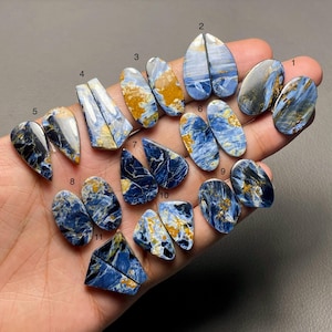 Puede incluir: Una colección de piedras preciosas de pietersita pulidas en varias formas, incluyendo ovaladas, de lágrima y rectangulares. Las piedras muestran una mezcla de colores azul, dorado y gris con patrones arremolinados únicos. Las piedras preciosas se sostienen en una mano.