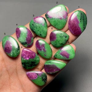 Puede incluir: Una colección de once piedras preciosas de rubí zoisita pulidas. Cada piedra muestra una mezcla de verde vibrante y magenta intenso, con patrones y formas únicos. Las piedras preciosas se sostienen en una mano, mostrando su belleza natural.