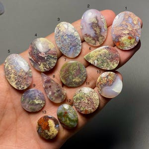 Pode incluir: Uma coleção de treze cabochões de pedras preciosas polidas em várias formas, incluindo ovais, em forma de gota e redondas. As pedras exibem uma mistura de cores, como vermelho, verde, castanho e branco, com padrões e texturas únicos. As pedras preciosas são seguradas em uma mão.