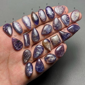 May include: A collection of 24 polished gemstones in various shapes, including oval, teardrop, and rectangular. The stones display a mix of purple, silver, and gold colors, with a sparkling, iridescent effect. The gemstones are numbered 1 through 24.