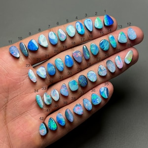 May include: A hand displays 42 polished opal gemstones in various shapes and sizes. The opals showcase a range of colors, including blues, greens, and whites, with visible internal patterns. Each stone is numbered for identification.