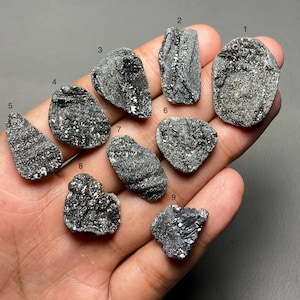 Può includere: Una collezione di nove pietre di pirite grezze, grigio scuro, ciascuna con una forma e una consistenza uniche. Le pietre sono tenute in una mano, mostrando il loro aspetto naturale e non lucidato. I cristalli di pirite hanno una lucentezza metallica.