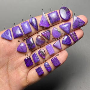 May include: A collection of polished purple sugilite gemstones in various shapes, including oval, square, triangle, and teardrop. The gemstones are displayed on a hand, showcasing their rich color and unique patterns. The image is well-lit, highlighting the details of each stone.