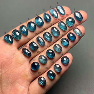 Op de afbeelding: Een verzameling van zevenentwintig blauwgroene edelstenen in verschillende vormen en maten, genummerd van 1 tot 27. De edelstenen zijn ovaal, druppelvormig en rond, met verschillende gradaties van transparantie. De edelstenen worden in een hand tegen een donkere achtergrond gehouden.