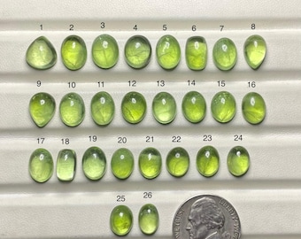 May include: A collection of 26 peridot gemstones in various shapes and sizes, ranging from oval to pear-shaped. The gemstones are a vibrant, translucent green color. A US nickel is included for size comparison.