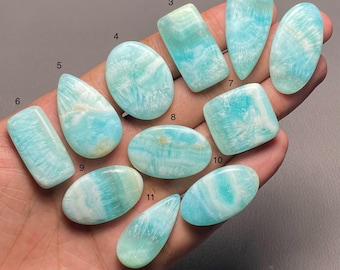 May include: A collection of eleven polished Amazonite gemstones in various shapes, including oval, rectangular, and teardrop. The stones display a mix of light blue and white colors with unique patterns. The gemstones are held in a hand.