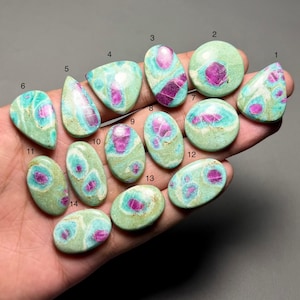 May include: A collection of fourteen polished gemstones, each with a unique shape and pattern. The stones feature a mix of colors, including teal, light green, and vibrant magenta. The gemstones are held in a hand against a neutral background.