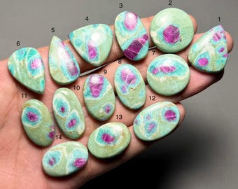 Può includere: Una collezione di quattordici pietre preziose lucide, ognuna con una forma e un motivo unici. Le pietre presentano una miscela di colori, tra cui verde acqua, verde chiaro e magenta vibrante. Le pietre preziose sono tenute in mano su uno sfondo neutro.