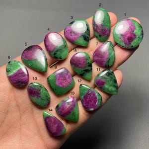 Puede incluir: Una colección de catorce piedras preciosas de rubí zoisita pulidas. Cada piedra muestra una vibrante mezcla de tonos magenta intenso y verde. Las piedras preciosas varían en forma, incluyendo ovalada, de lágrima y cuadrada.