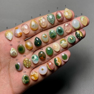 Op de afbeelding: Een verzameling van 33 gepolijste edelsteen cabochons in verschillende vormen en kleuren, waaronder groen, bruin, geel en roze. Elke steen is genummerd en wordt op een hand getoond, wat de verscheidenheid aan natuurlijke steenpatronen en -tinten laat zien.