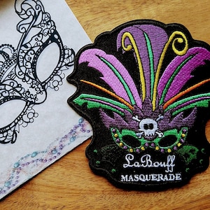 May include: A black and white embroidered mask with a crown and intricate details. A purple, green, and yellow embroidered patch with the text "La Bouff Masquerade" is also shown.