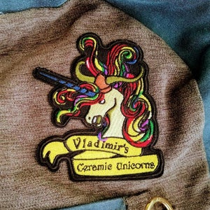 May include: A colorful embroidered patch featuring a unicorn with a rainbow mane and a yellow banner that reads "Vladimir's Ceramic Unicorns".