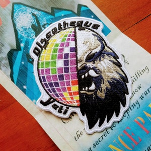 May include: A white embroidered patch with a disco ball and a wolf's head. The text "Discotheque Yeti" is embroidered around the edge of the patch.