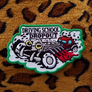 May include: A white and green embroidered patch with a cartoon image of a car driving off a cliff. The car is black and gray with red flames coming out of the back. The text "Driving School Dropout" is above the car.