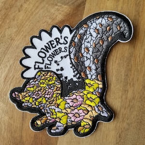 May include: A white, black, and yellow embroidered patch with a floral design and the text "FLOWER'S FLOWERS".
