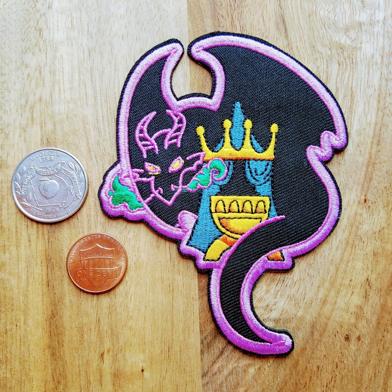 May include: A black and purple embroidered patch of a dragon with a crown and a goblet. The dragon is holding a goblet in its claws. The patch is on a wooden surface.