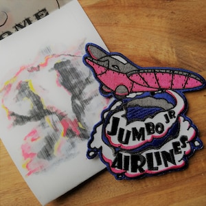 May include: Embroidered patch featuring a cartoon airplane with pink and gray details, flying through a white cloud with blue and pink lettering that reads "Jumbo Jr Airlines".