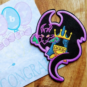 May include: Purple and black embroidered patch of a dragon wearing a gold crown. The dragon is holding a golden goblet in its claws. The patch has a green and yellow design.
