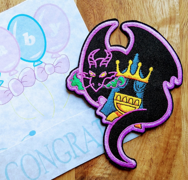 May include: Purple and black embroidered patch of a dragon wearing a gold crown. The dragon is holding a golden goblet in its claws. The patch has a green and yellow design.