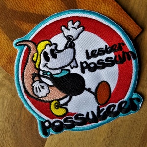 May include: A white, red, and blue embroidered patch with a cartoon possum holding a saxophone. The text "lesker possum" is written in a circle around the possum. The text "possum" is written in a larger font at the bottom of the patch.