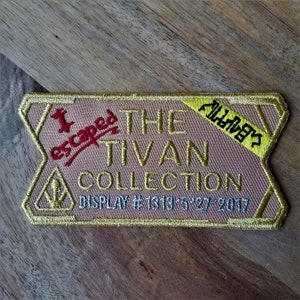 May include: A gold and brown embroidered patch with the text "I escaped THE TIVAN COLLECTION DISPLAY #1319 5 27 2017" and a yellow tag with the text "All rights reserved".